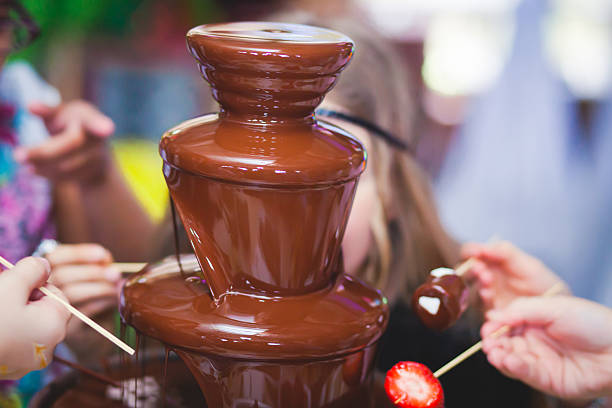 A chocolate fountain with hands reaching for strawberries and other treats.