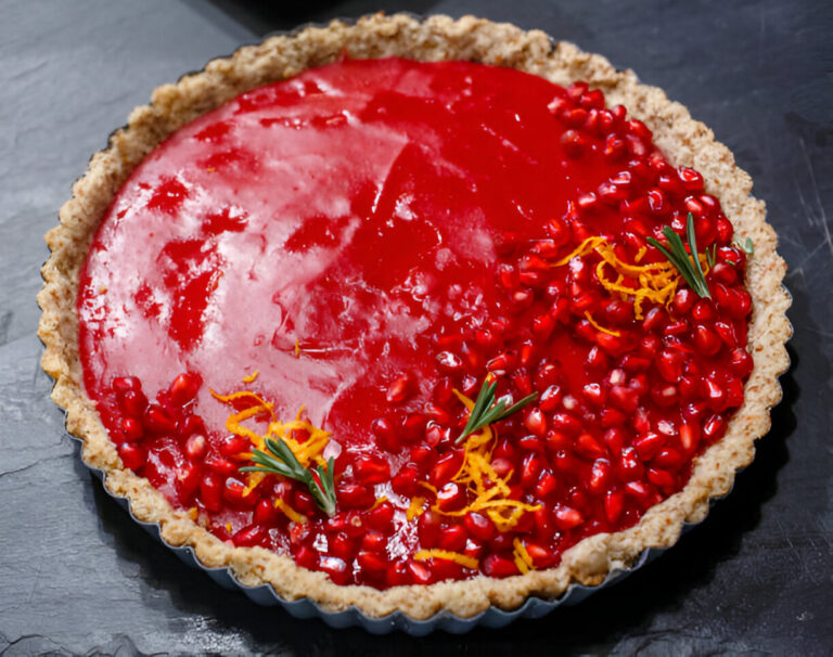 A vibrant pomegranate tart with a glossy red filling topped with pomegranate seeds, orange zest, and rosemary sprigs on a dark surface.