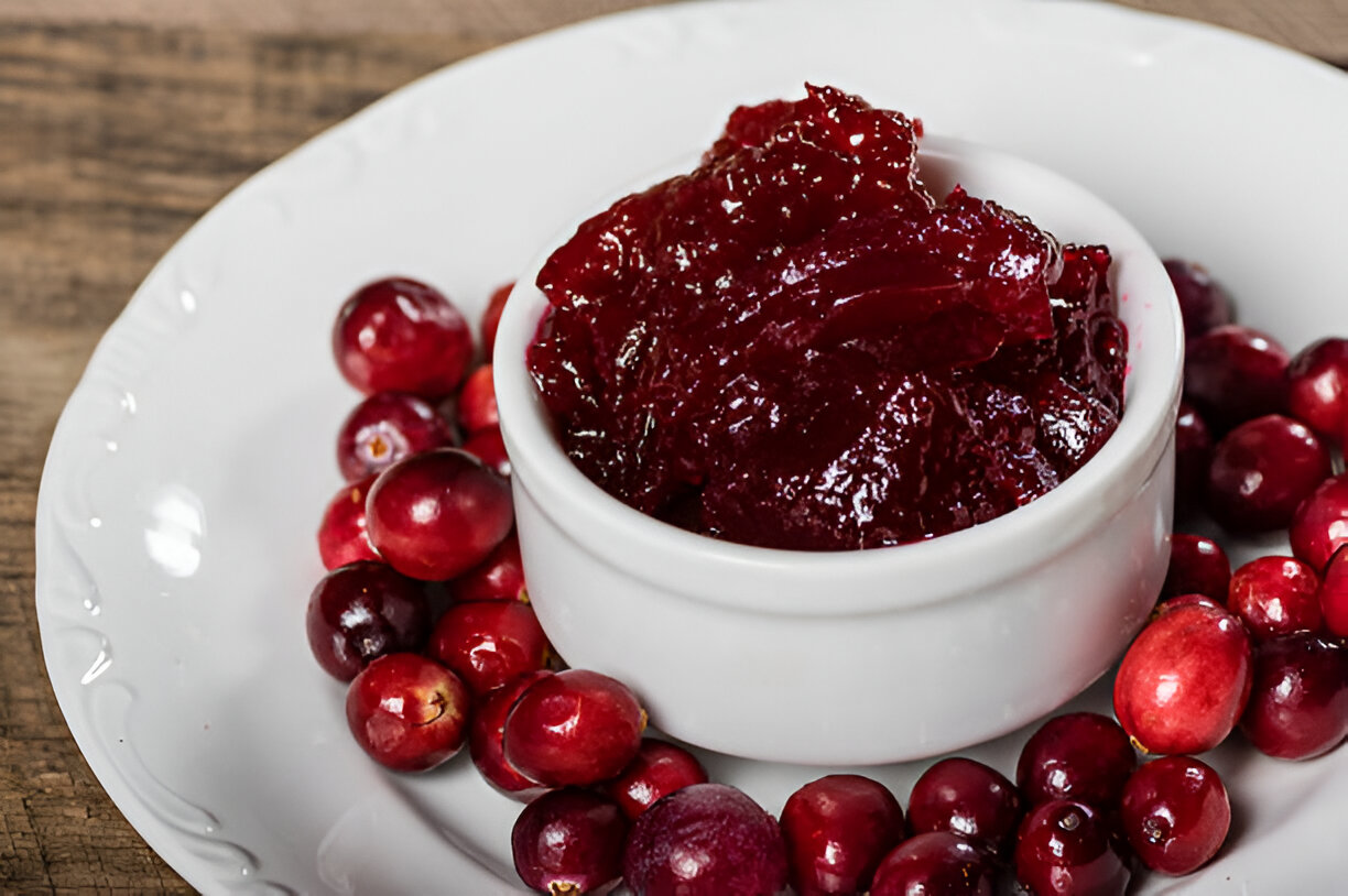 A white dish filled with cranberry sauce surrounded by fresh cranberries on a wooden surface.