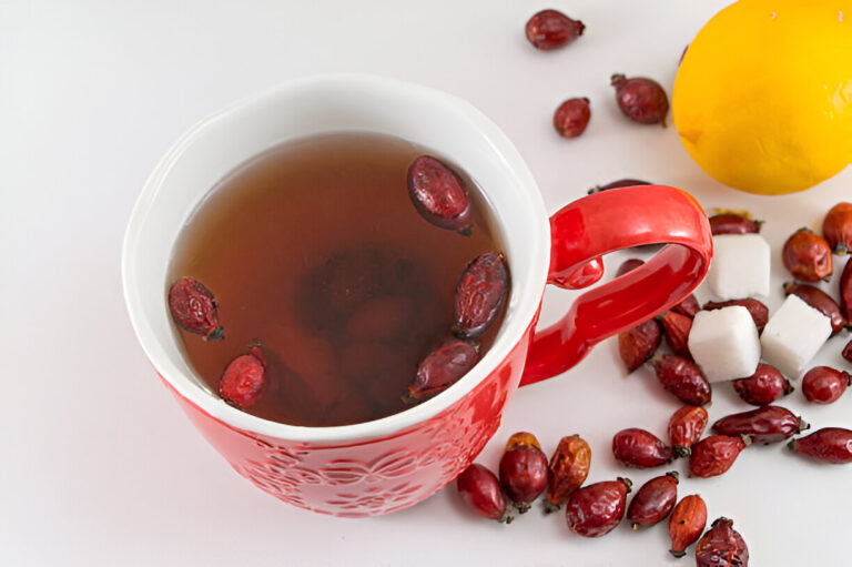 A cup of herbal tea with rose hips, a yellow lemon, and sugar cubes on a white surface.