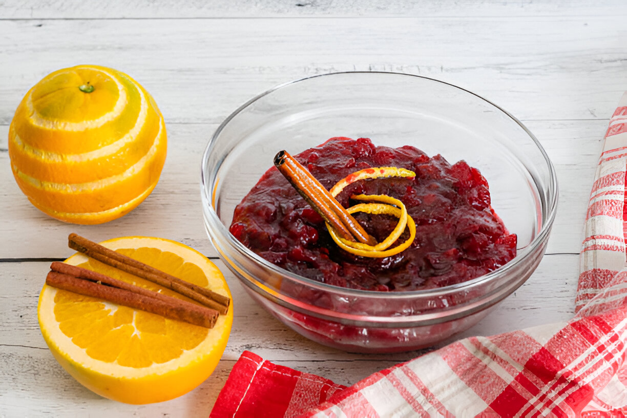 A bowl of cranberry sauce garnished with cinnamon sticks and citrus zest, alongside a halved orange and a red checkered cloth.