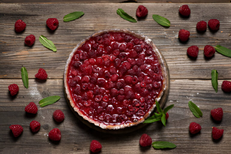 Delicious raspberry pie surrounded by fresh raspberries and mint leaves on a wooden table.