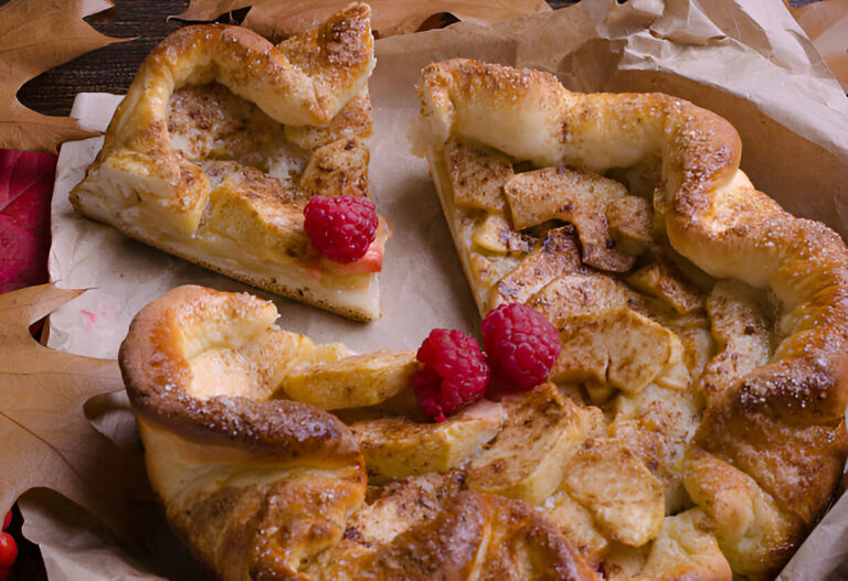 A delicious apple tart with raspberry garnishes on a parchment-lined surface surrounded by autumn leaves.