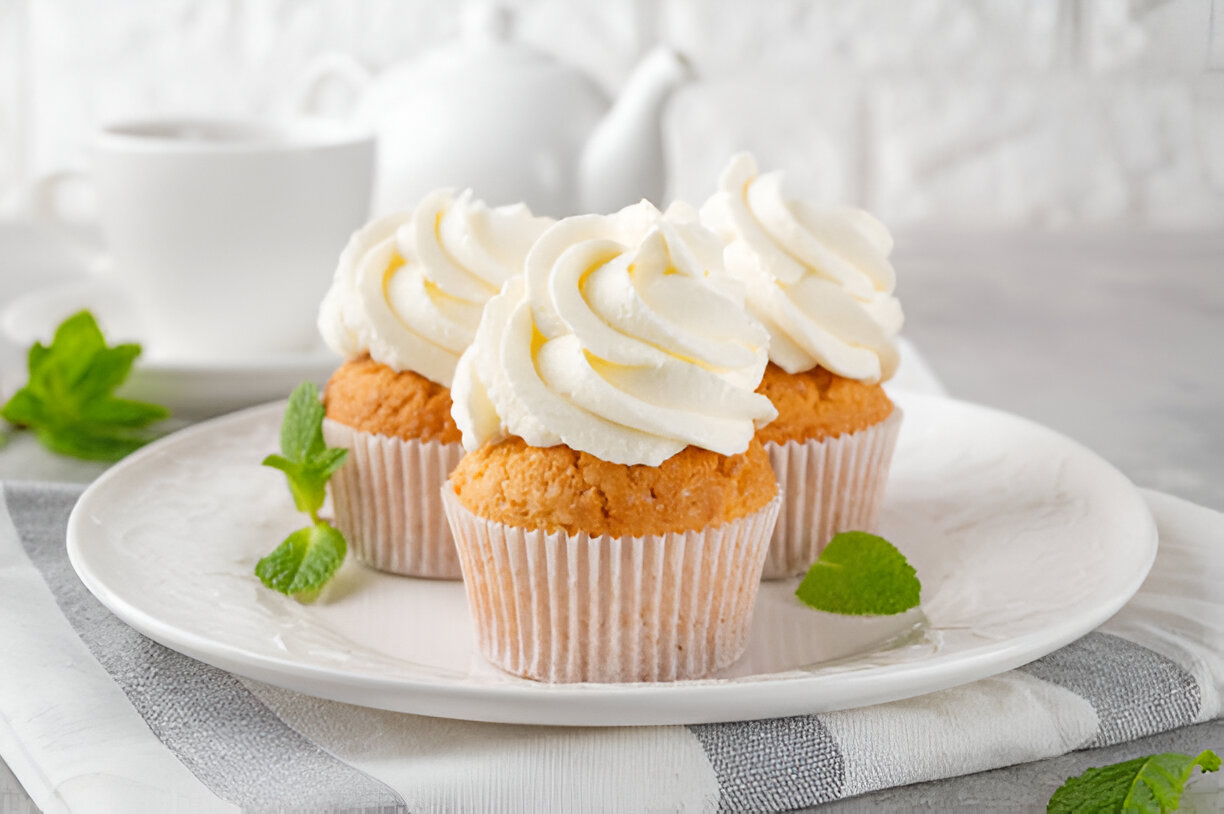 A close-up of two fluffy cupcakes topped with creamy white frosting, placed on a white plate with mint leaves.