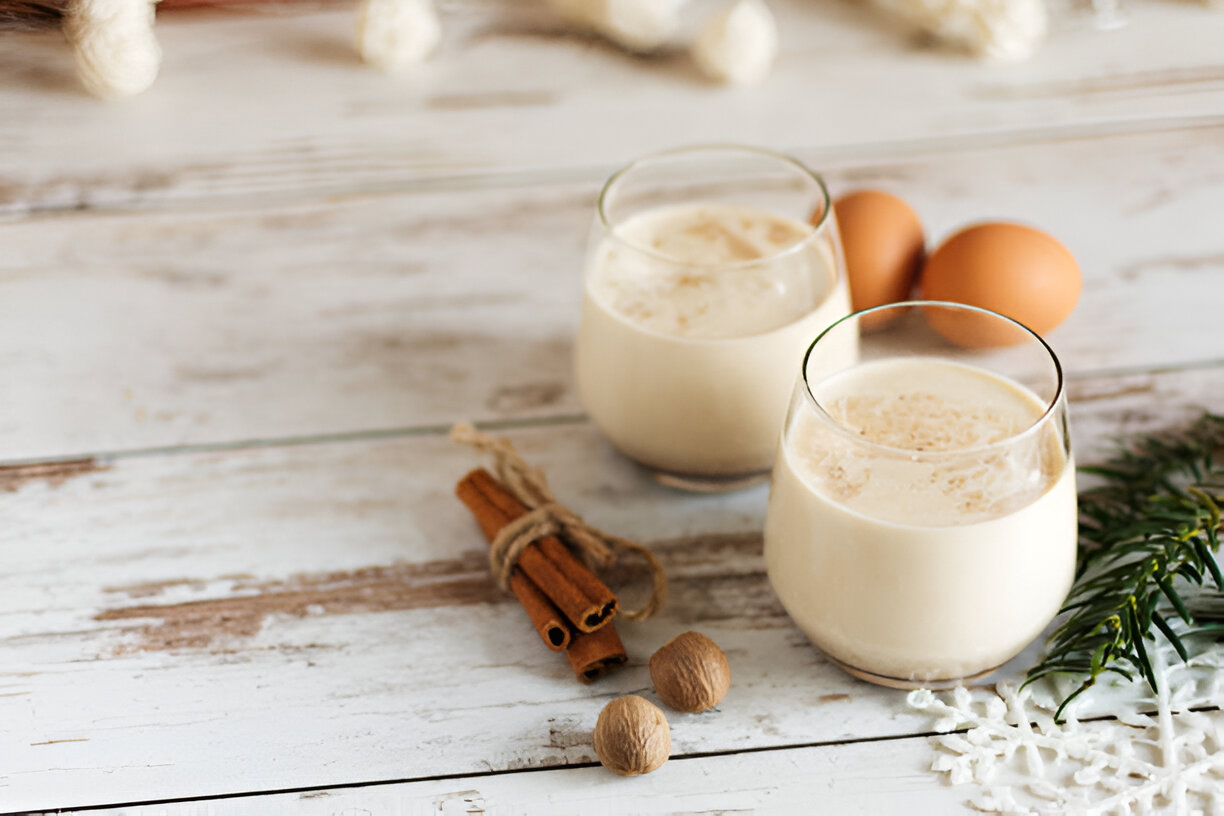 Two glasses of creamy eggnog with cinnamon sticks, eggs, and nutmeg on a rustic wooden table.