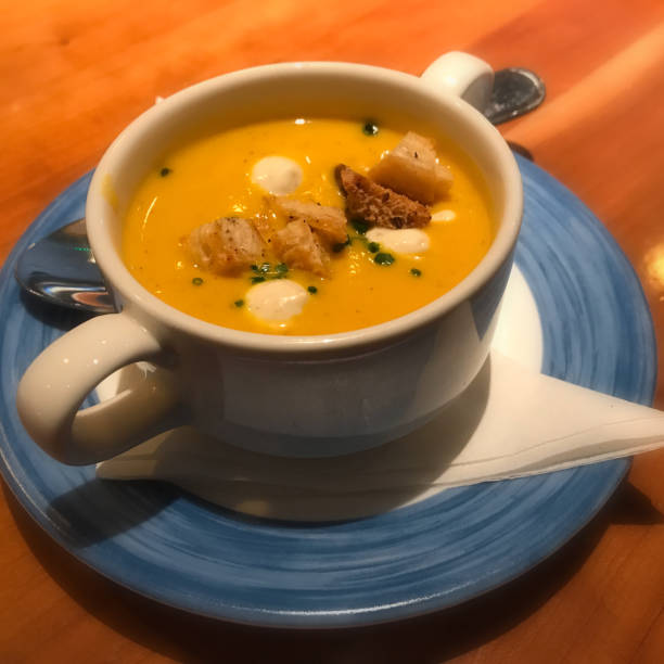 Cup of vibrant yellow soup garnished with croutons and cream on a blue plate.