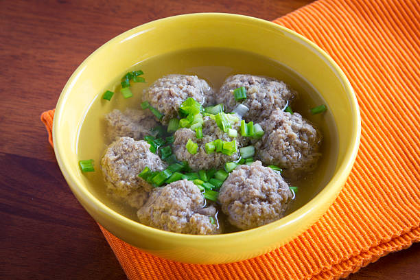 A bowl of meatballs in clear broth, garnished with chopped green onions, placed on a textured orange napkin.