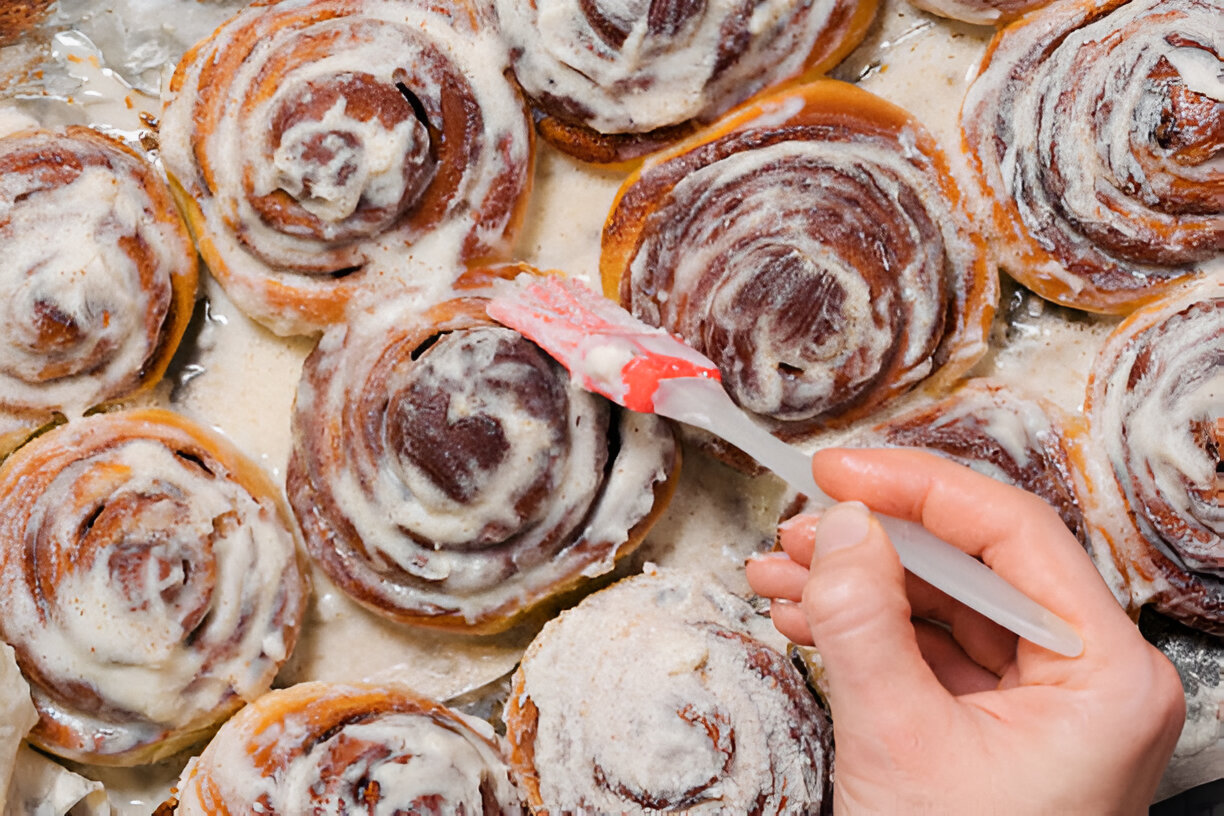 A hand applying icing to freshly baked cinnamon rolls.