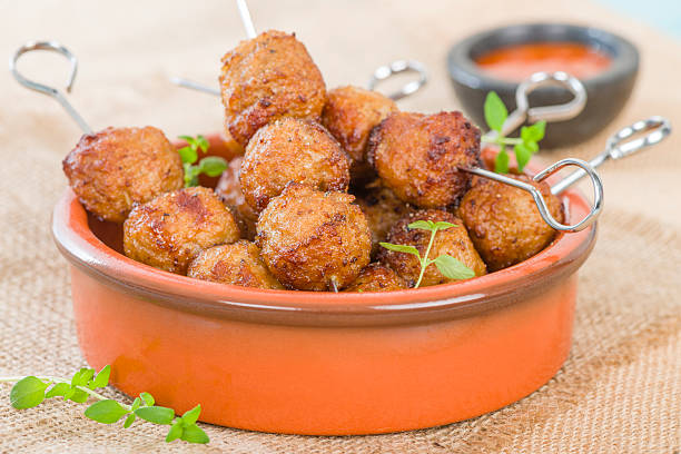 A close-up of crispy golden-brown meatballs served in an orange bowl with skewers, garnished with green herbs, alongside a small bowl of dipping sauce.