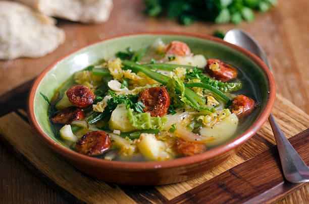 A bowl of hearty soup with vegetables and sliced sausage, garnished with fresh herbs, served on a wooden cutting board.