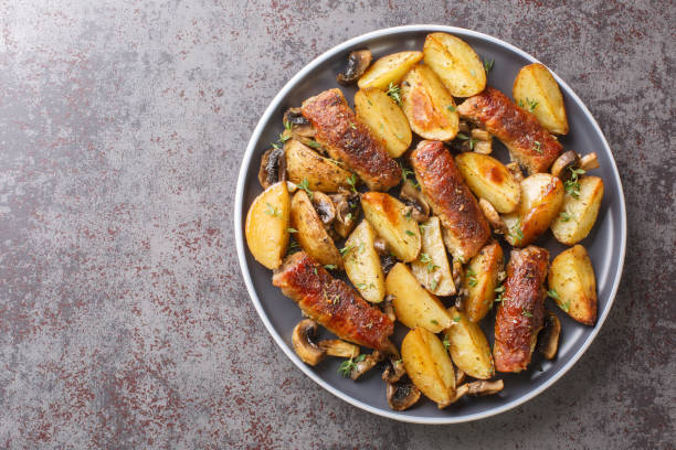 A plate of roasted sausages with golden-brown potatoes and mushrooms arranged on a textured surface.