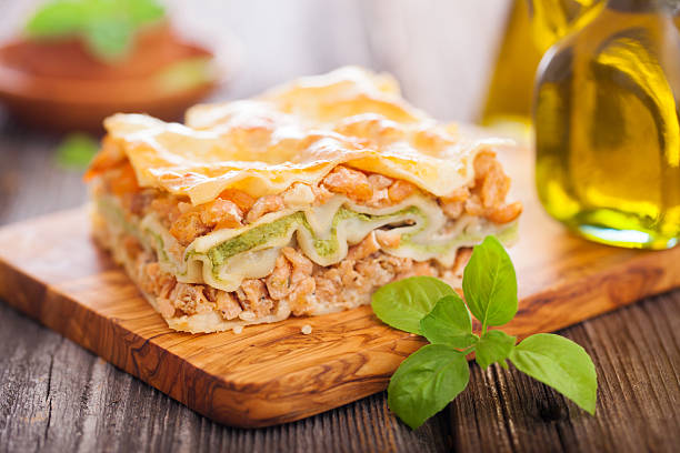 A slice of lasagna on a wooden cutting board, garnished with fresh basil leaves and accompanied by a bottle of olive oil.