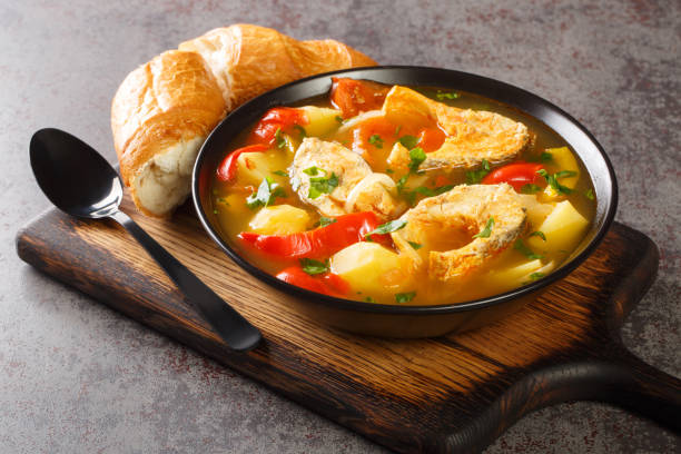 A bowl of fish soup with vegetables served on a wooden cutting board alongside a croissant and a spoon.