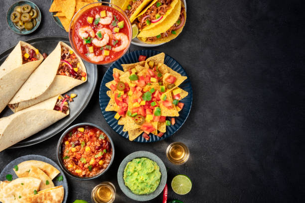 A colorful spread of Mexican cuisine featuring nachos, tacos, guacamole, salsa, and tortillas on a dark surface.