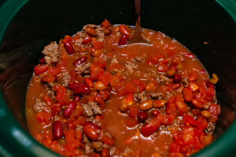 A close-up of a hearty chili dish simmering in a green crockpot, featuring ground meat, kidney beans, diced tomatoes, and various spices.