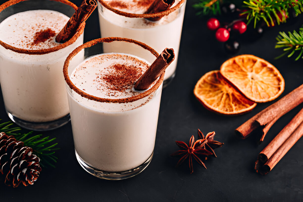 Three cocktails with a creamy texture, garnished with cinnamon sticks and topped with chocolate powder, alongside dried orange slices and pinecones on a dark surface.