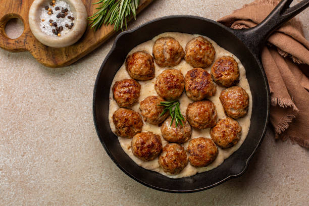 A skillet filled with meatballs in a creamy sauce, garnished with rosemary, accompanied by a side of salt and pepper.