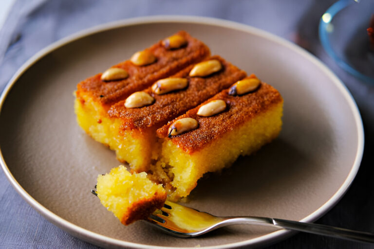 A piece of golden cake topped with whole almonds on a gray plate, with a fork and a partially consumed piece of cake.