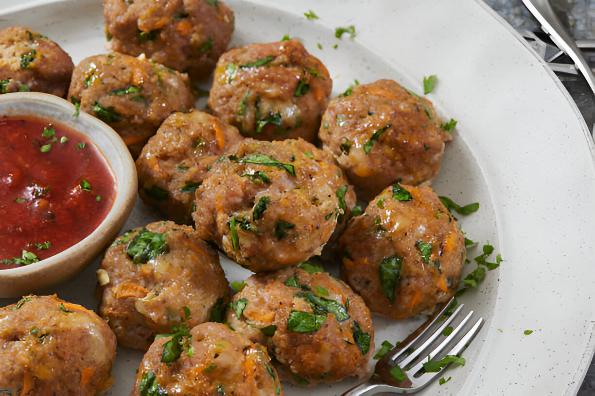 A plate of meatballs garnished with fresh herbs, served with a small bowl of red sauce.