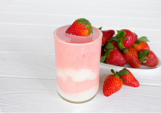 A refreshing layered strawberry smoothie in a glass, topped with a fresh strawberry, with a plate of strawberries in the background.