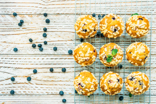 A cooling rack with freshly baked muffins topped with crumb and garnished with mint leaves, surrounded by scattered blueberries on a wooden surface.