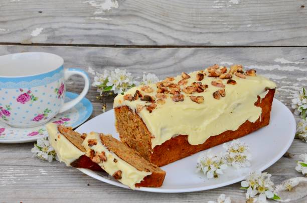 A loaf of cake topped with frosting and walnuts, served with a cup of tea on a wooden table.