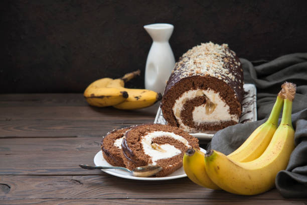 Chocolate roll cake with banana filling on a wooden table, accompanied by bananas and a white jug.