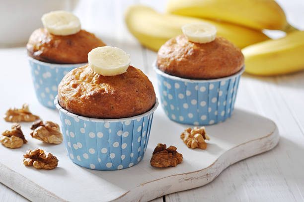 Three freshly baked banana muffins topped with banana slices in blue polka dot liners, surrounded by walnuts and bananas in the background.