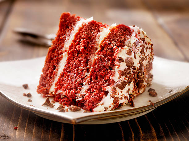 A slice of red velvet cake topped with cream cheese frosting and chocolate shavings on a white plate.