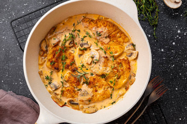 A creamy chicken dish with mushrooms and thyme in a large white skillet, served on a dark textured surface.