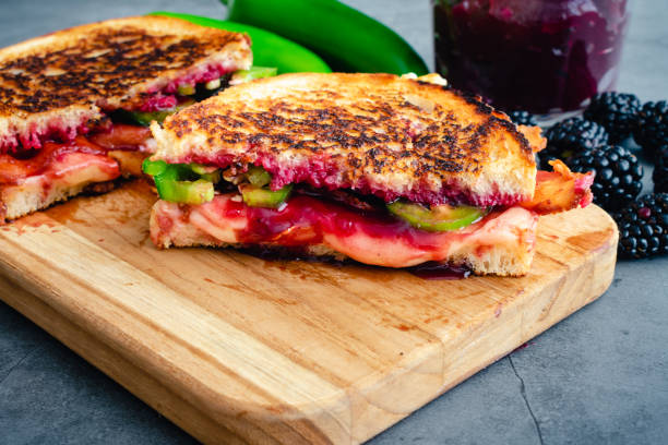 A close-up of a grilled sandwich with mixed fillings including jelly and green peppers, served on a wooden cutting board with blackberries and a jar in the background.