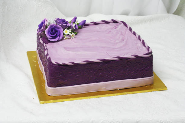 A beautifully decorated square cake with purple frosting and rose decorations on top, placed on a gold cake board.