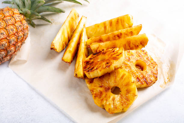 Grilled pineapple slices and spears on a parchment paper with a fresh pineapple in the background.