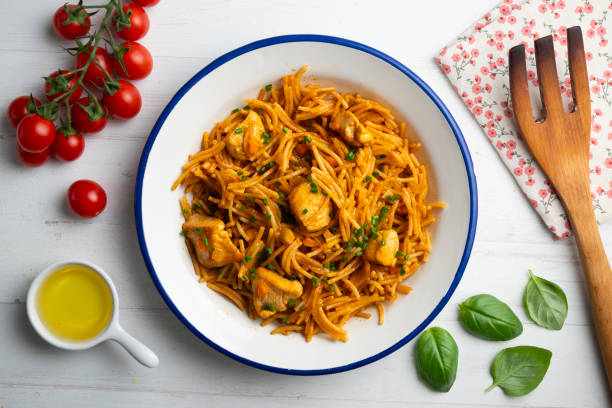 A bowl of pasta with chicken pieces, garnished with herbs, placed on a white wooden table with cherry tomatoes, olive oil, and a wooden spatula in the background.