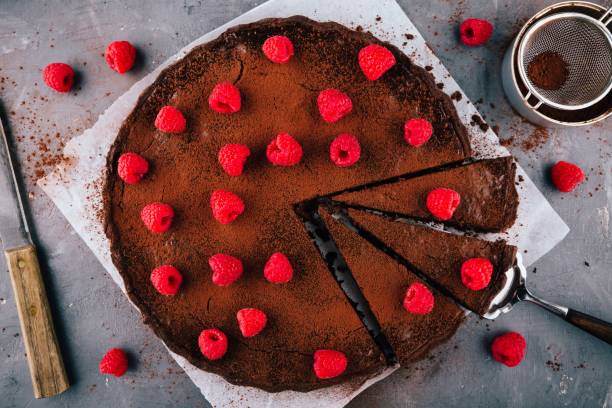 A chocolate tart topped with fresh raspberries and cocoa powder, with a slice cut out and a serving spatula beside it.