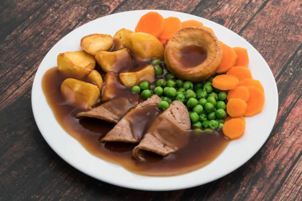 A plate of roast beef with gravy, roast potatoes, green peas, sliced carrots, and Yorkshire pudding.