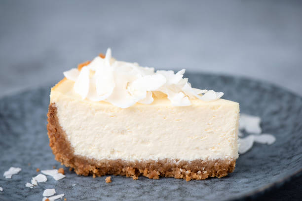 A slice of creamy cheesecake topped with shaved coconut on a dark plate.