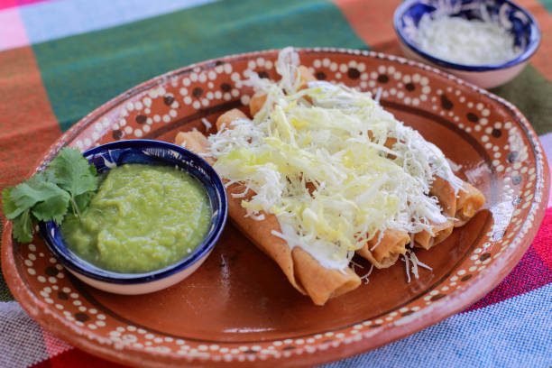 A traditional dish of flautas topped with shredded cheese served on a colorful tablecloth with a side of green sauce.