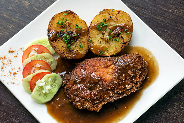 A plate of fried chicken with gravy, served with two baked potatoes and a side of fresh sliced vegetables.