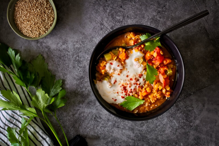 A bowl of colorful vegetable stew topped with yogurt and spices, alongside fresh parsley and sesame seeds.