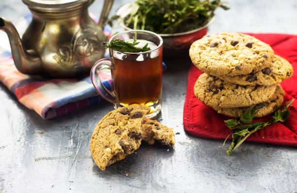 A glass of tea with mint leaves alongside a stack of chocolate chip cookies and a teapot.