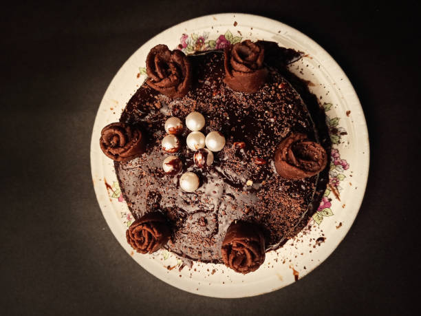 A beautifully decorated chocolate cake with chocolate rose decorations and white pearls on a floral plate against a dark background.