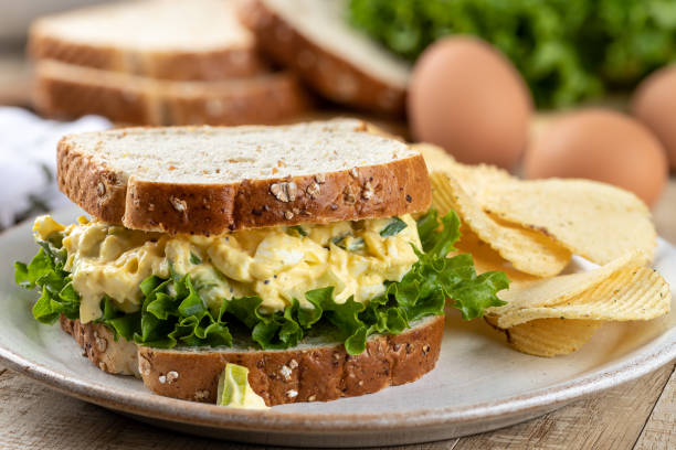 A sandwich made with oats bread filled with egg salad and lettuce, accompanied by a side of potato chips and fresh eggs.