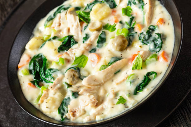 A creamy chicken and vegetable stew in a black bowl, featuring spinach, gnocchi, and colorful vegetables.