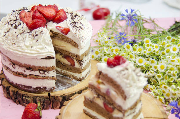 A beautifully layered chocolate and strawberry cake with sliced pieces on a wooden board, surrounded by flowers.