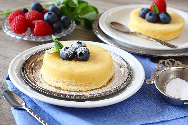 Two delicious yellow cake desserts topped with blueberries and mint, served on silver plates with fresh raspberries in the background.