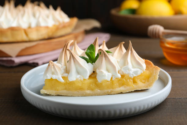 A slice of lemon meringue pie on a white plate with mint leaves, set against a dark wooden background.