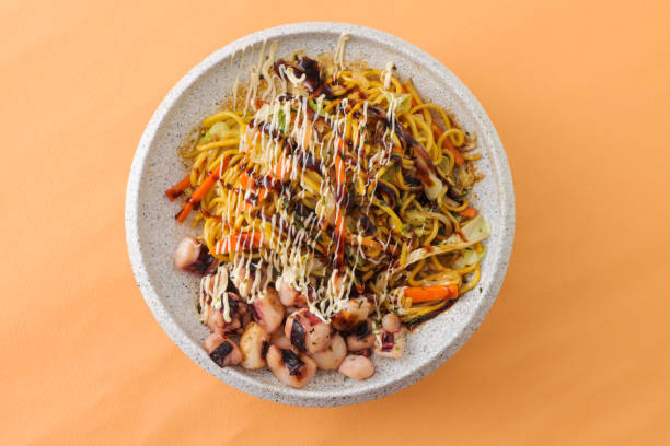 A bowl of fried noodles topped with colorful vegetables and drizzled with sauce, served on an orange background.