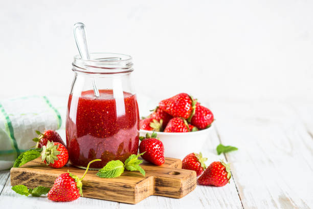 A jar of strawberry jam on a wooden cutting board, surrounded by fresh strawberries and mint leaves.