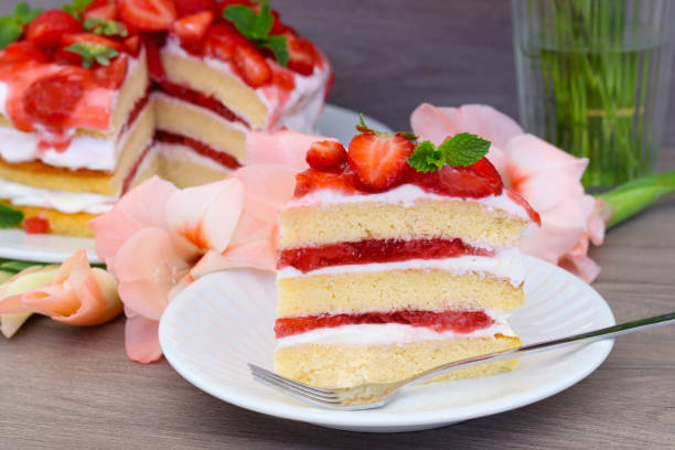 A slice of strawberry cake with layers of cream and strawberries, garnished with mint leaves, on a white plate next to a whole cake and pink flowers.